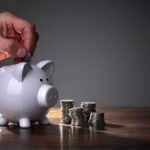 Brain Hack Tricks Americans Into Saving Billions A hand inserting a coin into a white piggy bank on a wooden table with stacks of coins nearby