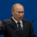 Putin FIRES Advisor Who Supports Peace A man in a suit gesturing while speaking at a podium with microphones against a blue background