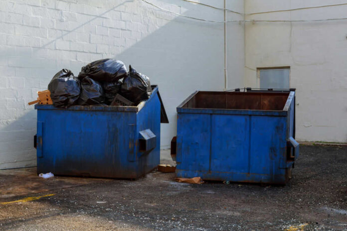 Two blue dumpsters, one overflowing with trash bags.