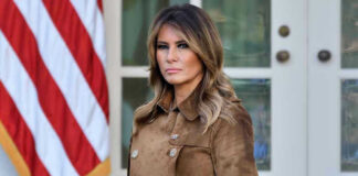 Woman in brown coat beside American flag.
