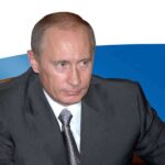 A serious-looking man in a suit sitting at a table with a Russian flag in the background