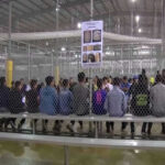 New ICE Detention Center REVEALED – 1,500 Beds! People sitting on benches inside a fenced facility.