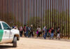 Children Separated Twice After Trump Crackdown! People walking beside tall fence and border patrol vehicle.