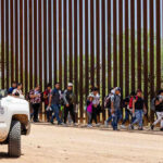 Children Separated Twice After Trump Crackdown! People walking beside tall fence and border patrol vehicle.