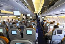 Mid-Flight Mayhem – Passengers TACKLE Maniac! Interior view of an airplane with passengers seated and using in-flight entertainment screens
