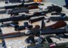 DOJ Slams Blue-Cities $1,591 Gun Fee Assorted firearms and ammunition laid out on a wooden table