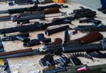 DOJ Slams Blue-Cities $1,591 Gun Fee Assorted firearms and ammunition laid out on a wooden table