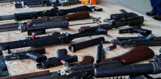 DOJ Slams Blue-Cities $1,591 Gun Fee Assorted firearms and ammunition laid out on a wooden table