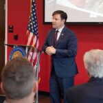 Vanished Congressman RETURNS – Been Missing For MONTHS! Speaker in a navy suit addressing an audience at a podium