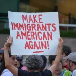 Washed-Up John Kasich Defies Court – Why Now? Teddy Williams at a protest holding a sign reading 'Make Immigrants American Again'