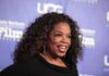 Oprah Tells Audience To CELEBRATE Murderous Mothers smiling woman with curly hair on a blue step-and-repeat backdrop