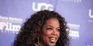 Oprah Tells Audience To CELEBRATE Murderous Mothers smiling woman with curly hair on a blue step-and-repeat backdrop