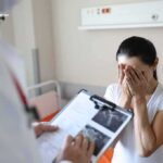 Teen Movie Icon Passes Away – Cause Revealed Woman covering her face while doctor holds a clipboard in a hospital room