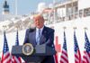 Trump Offers Pathetic Excuse For Rising Cost of Living Donald Trump speaking at a podium with American flags and a ship in the background