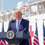 Trump Offers Pathetic Excuse For Rising Cost of Living Donald Trump speaking at a podium with American flags and a ship in the background