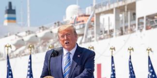 Trump Offers Pathetic Excuse For Rising Cost of Living Donald Trump speaking at a podium with American flags and a ship in the background