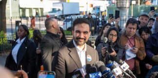 Zohran Mamdani speaks at a press conference; Julia Bacha is visible among the crowd