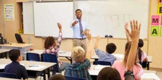 Officials Warn Parents Of Unsettling Viral Trend Affecting Kids Teacher at front of classroom as students raise hands