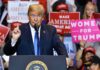 Donald Trump speaking at a rally podium with crowd holding signs