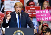 Donald Trump speaking at a rally podium with crowd holding signs