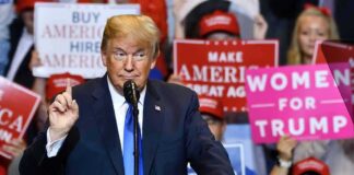 Donald Trump speaking at a rally podium with crowd holding signs