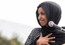Ilhan Omar speaking at a microphone with hand on chest