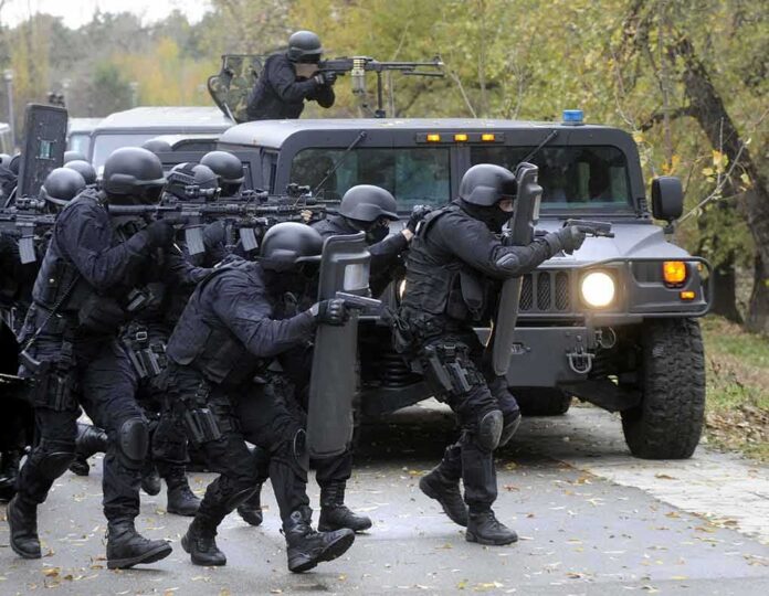 shutterstock_346794716 Tactical police team advances from armored vehicle with shields and rifles